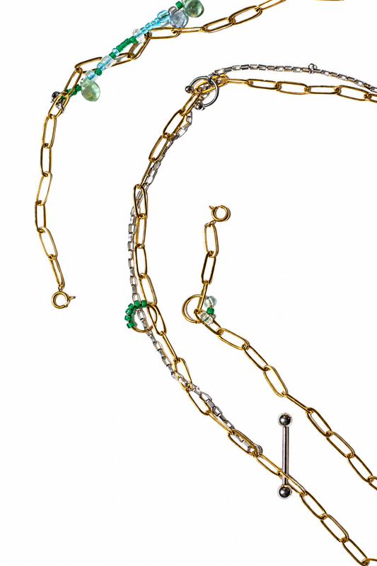 Gros plan sur une chaîne de ventre en acier inoxydable plaqué PVD or et acier inoxydable argent avec perles vertes et bleues, anneaux décoratifs et détails inspirés du piercing.