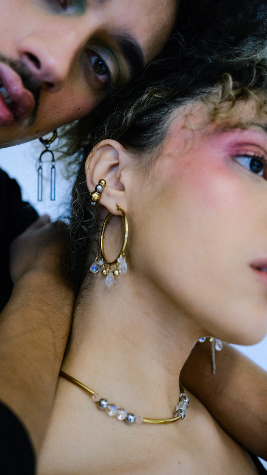 Close-up of a woman wearing gold hoop earrings and a necklace, with a man's face partially visible.
