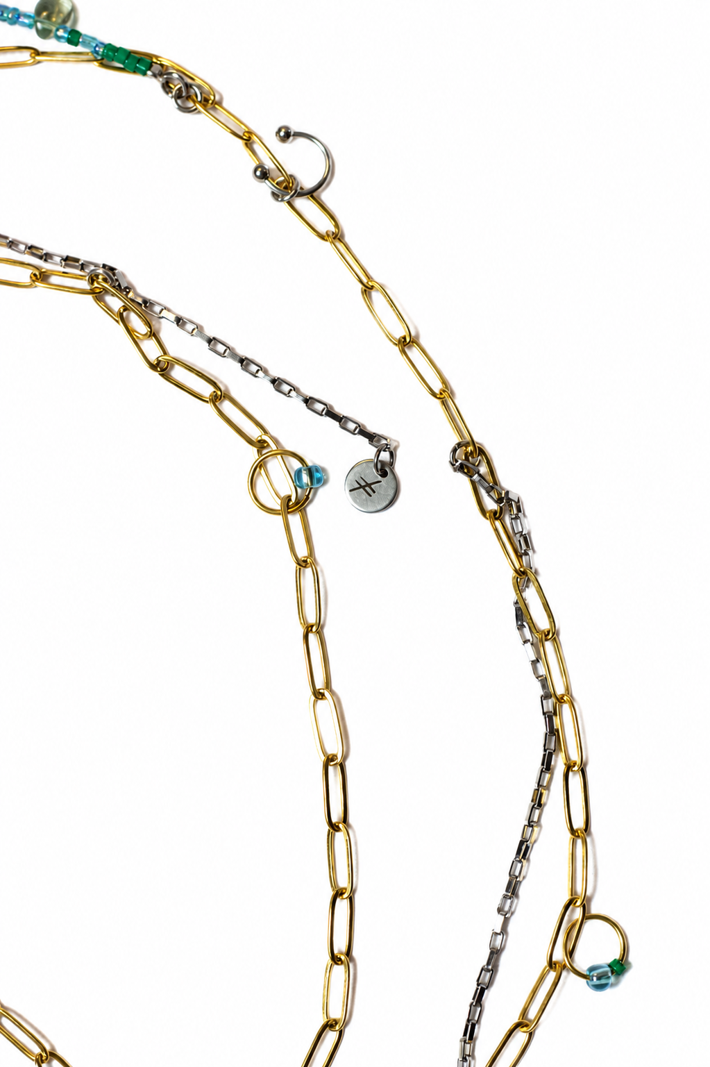 Chaîne de ventre en acier inoxydable or et argent avec fermoirs aux extrémités, détails verts et bleus et éléments inspirés du piercing, vue à plat sur fond blanc.