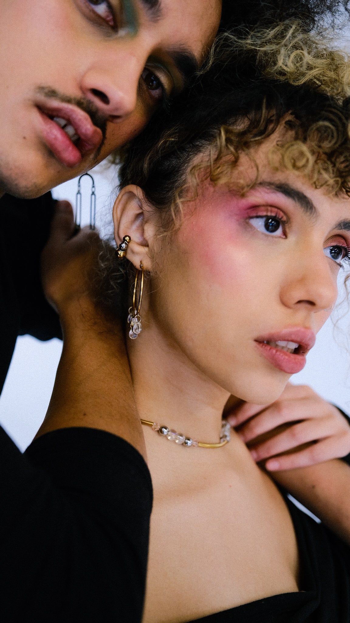 Two people close together, one with curly hair and makeup, wearing jewelry.