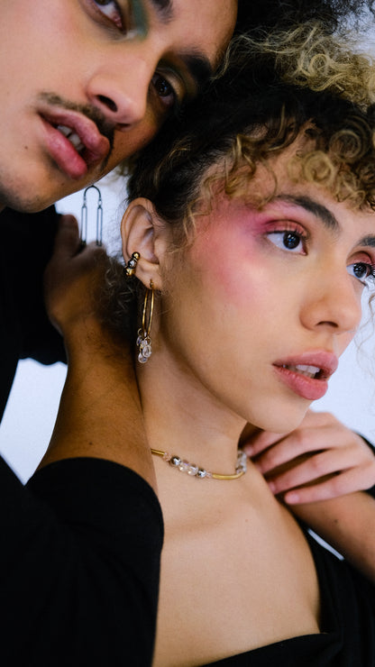 Two people close together, one with curly hair and makeup, wearing jewelry.