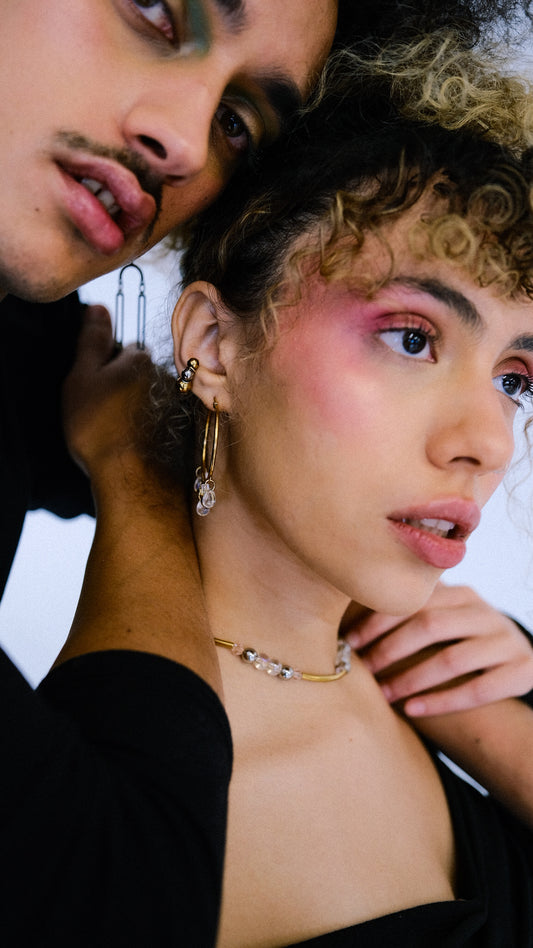 Two people close together, one with curly hair and makeup, wearing jewelry.