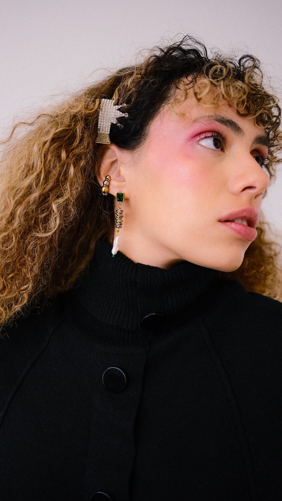 Woman with curly hair wearing a black coat and earrings against a neutral background